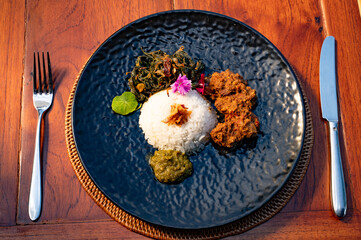 A dark plate of Indonesian food with rice, meat, and vegetables, ready to be eaten.