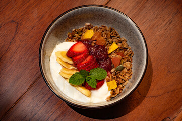 Overhead shot of a bowl filled with granola, yogurt, sliced strawberries, bananas, jam, and mint on a wooden surface.