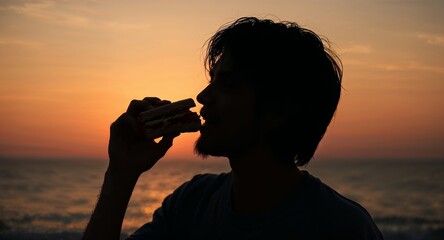 Silhouette of a man eating a sandwich against an orange sky and ocean at sunset or sunrise