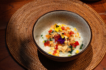 A bowl of rice porridge topped with colorful vegetables and edible flowers on a woven placemat.