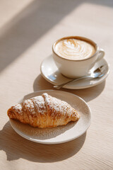 Cornetto pastry rests beside a creamy cappuccino in a white ceramic cup