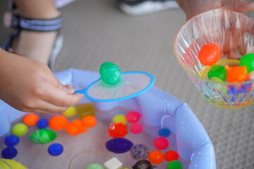 子供が屋外の水遊び用プールで鮮やかな緑色のスーパーボールを網に乗せ、横にある器に移そうとしている瞬間