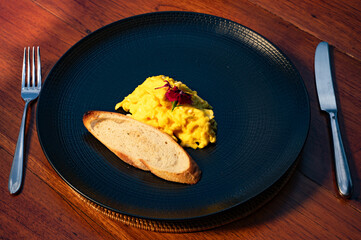 Scrambled eggs, bread, and cutlery arranged on a dark plate.