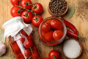 Tasty pickled tomatoes in jars and ingredients on wooden table, flat lay