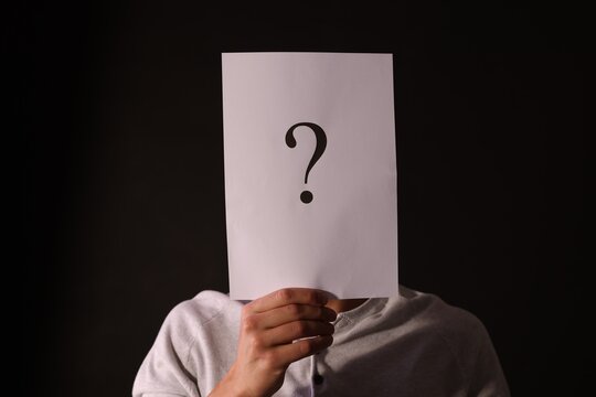 Man hiding behind paper sheet with question mark on dark background