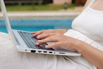 
A person's hands are busy typing on a silver laptop, with a swimming pool and blue water visible...