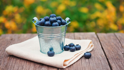 Metal bucket filled with blueberries stands on light napkin on wooden surface. Calm summer background.