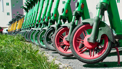 A long line of green electric scooters with red wheels parked on asphalt beside a grassy verge, illustrating urban dockless rental micromobility Photo