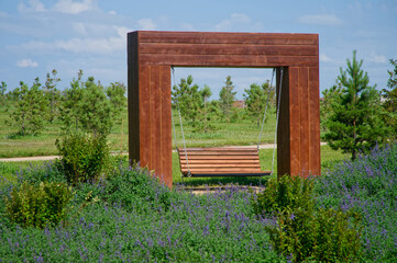 A wooden swing bench suspended within a minimalist timber frame, surrounded by purple flowering groundcover and young trees in a city park Photo