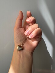 Female hand holding gold heart pendant necklace against neutral background. Elegant jewelry concept with soft light and shadows.