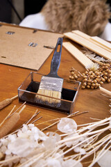 Close-up of a hand holding a brush dipped in black paint, surrounded by dried flowers and natural...