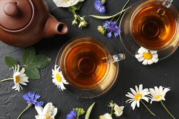 Aromatic herbal tea in glass cups, different flowers and teapot on black table, flat lay