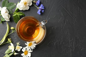 Aromatic herbal tea in glass cup, different flowers and mint on black table, flat lay. Space for...