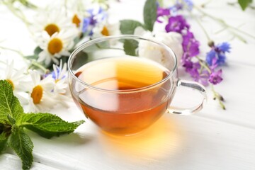 Aromatic herbal tea in glass cup, different flowers and mint on white wooden table, closeup