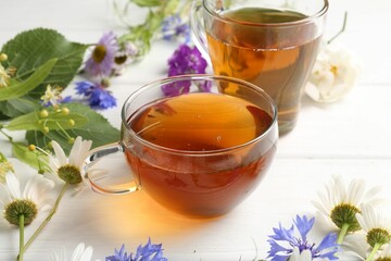 Aromatic herbal tea in glass cups, different flowers and green leaves on white wooden table, closeup