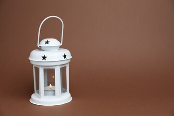 Beautiful white decorative lantern with burning candle on brown background, space for text