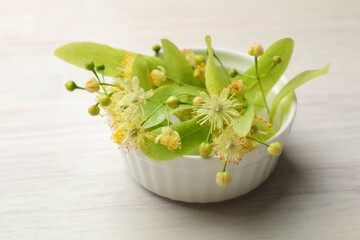 Beautiful linden flowers and leaves in bowl on white wooden table, closeup