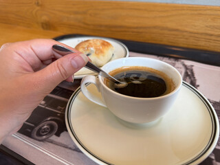Cup of black coffee being stirred by hand with a spoon. Small bun on a side plate in cozy cafe setting