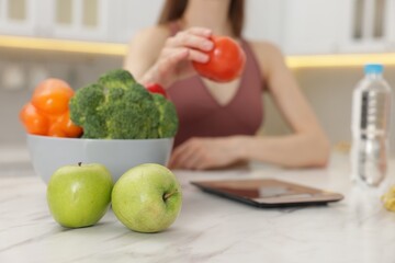 Woman weighting tomato on kitchen scale at white marble table indoors, selective focus