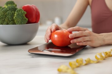 Woman weighting tomato on kitchen scale at white marble table indoors, closeup