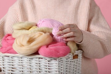 Woman holding wicker basket with colorful felting wool and yarns on pink background, closeup