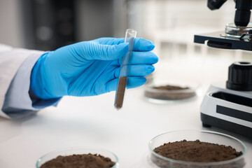 Scientist with test tube of soil sample at white table in laboratory, closeup