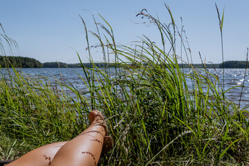 Relaxing Summer Day by the Lake — Female Legs in Grass Near Water with Scenic View and Bright...