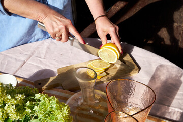 Close-up of a woman slicing a lemon on a wooden board. Sunlight highlights fresh citrus, glassware,...