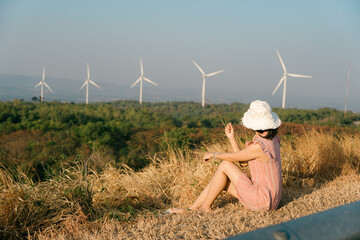 Young girl sitting on grassy hillside wea hat and pink dress looking at wind turbines in the...