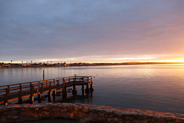 Fototapeta premium The sun rises above a calm bay, casting a golden glow over wooden picnic tables and tranquil waters, creating a perfect moment of stillness.