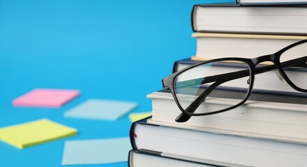 Detailed close-up of eyeglasses resting atop a pile of textbooks, set against a vibrant blue background scattered with colorful sticky notes, symbolizing study and organization