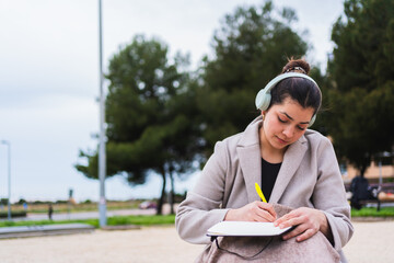 Young influencer taking notes on a notebook while listening to music with headphones in a park