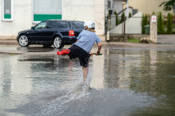 Boy riding scooter through urban floodwater in summer – fun, childhood and weather concept