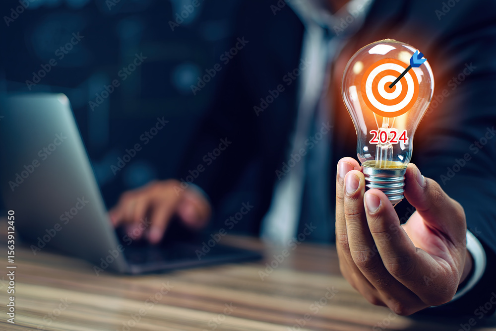 Poster illuminated light bulb with target and arrow displaying 2024, held by a person working on a laptop, 