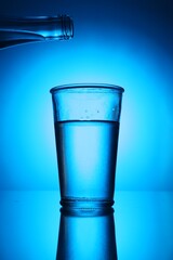 Bottle above glass of fresh water on blue gradient background. Backlit