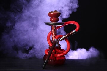 Hookah on wooden table with smoke in color lights against black background