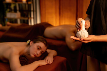 Hot herbal ball spa massage body treatment, masseur gently compresses herb bag on couple customer...