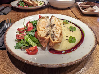 Fine dining meal: Beautifully plated baked fish with mashed potatoes, berry sauce, and a side salad.Delicious dinner plate featuring savory fish strudel, potato mash, and a salad with cherry tomatoes