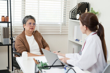 Fototapeta premium Elderly asian female sitting in wheelchair discusses symptoms of bone and joint pain with orthopedic doctor inside clinic office, having diagnostic consultation with notes and medicine bottle