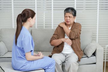 Young female physiotherapist listening while senior asian woman sitting on sofa explaining shoulder...