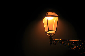 A classic street lamp emits a warm glow, casting light on a nearby surface, illuminating the area subtly in a dark environment.