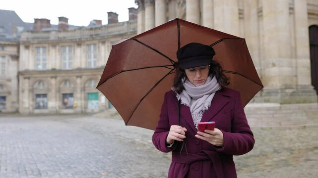 Young adult tourist woman holding an umbrella during rainy day using an app on her mobile phone to find a location in Paris city.