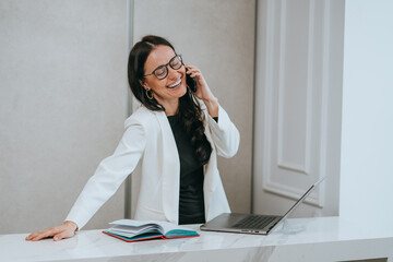 A joyful woman in a white blazer laughs while talking on the phone, embodying joy and connection in...