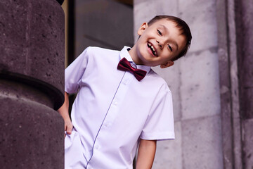 Cheerful schoolboy in white shirt and bow tie laughing and tilting from behind of pillar of school...