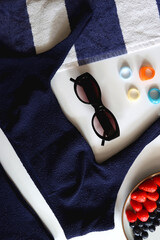 Navy swimsuit, striped beach towel, sunglasses, colorful rings and plate with berries. Top view.