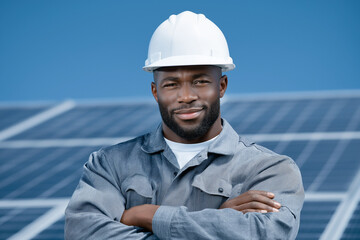 Construction worker stands with crossed arms at solar energy installation. Bright blue sky and solar panels create a vibrant backdrop. Concept of renewable energy, construction, sustainability