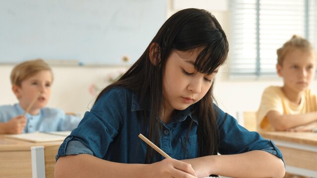Asian girl writing answer in paper while focus on thinking about theory during test. Diverse smart students taking a note or lecturing while teacher teaching in elementary class. Education. Pedagogy. - Powered by Adobe