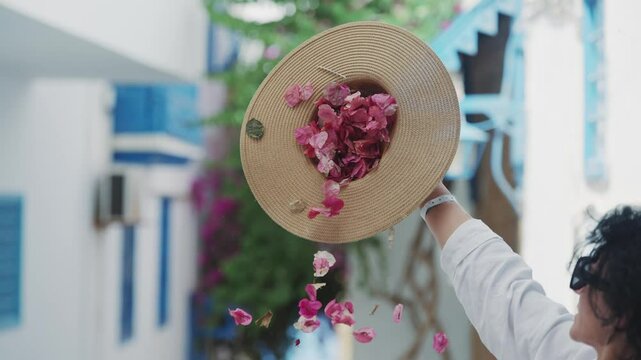 Woman pouring pink flowers from straw hat in Sidi Bou Said mediterranean village in Tunisia, slow motion