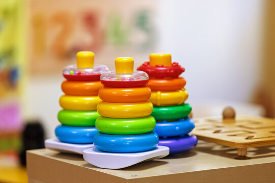 A trio of vibrant, colorful stacking ring toys stands on a wooden surface, ready for play in a bright kindergarten setting. The image evokes childhood learning and playful exploration.