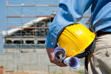 Construction worker holding yellow hard hat and blueprints at building site